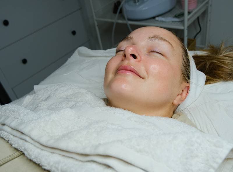 A woman with a content expression lies on a treatment bed covered with a white towel, receiving a facial skincare procedure in a clinic.