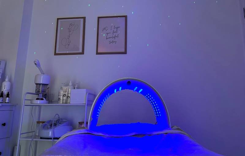 A serene beauty salon room with a glowing blue LED light therapy facial treatment bed.