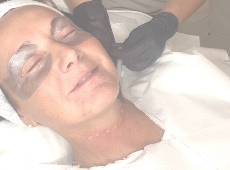 A woman receiving a Bio Nutri Neck Treatment with closed eyes, as a technician applies a mask to her face.