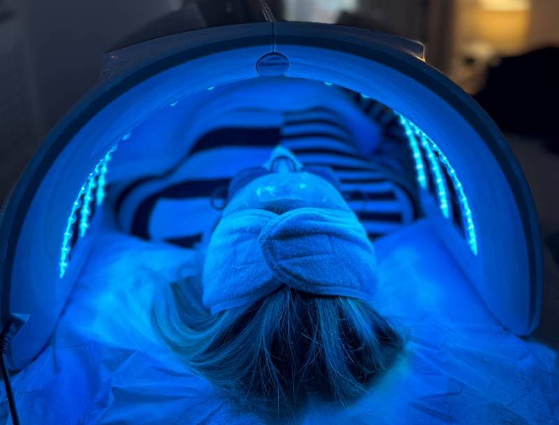 Person undergoing a facial treatment using blue led light therapy at Assured Aesthetics, lying inside a dome-shaped device.