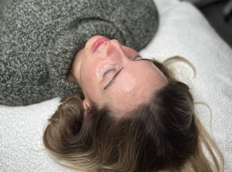 A woman lying on her back with her eyes closed, resting on a padded surface, receiving a facial, wearing a gray knitted sweater.