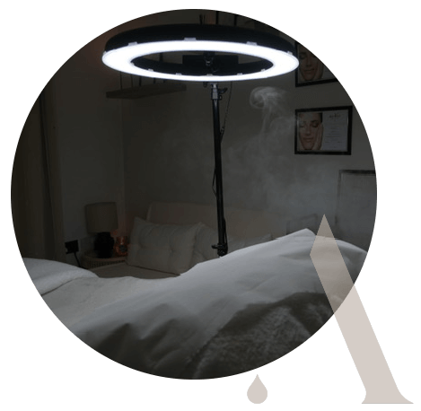 A circular light on a stand illuminates a white bed in a dimly lit room with soft furnishings and photographs on the wall.